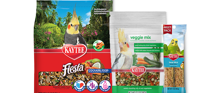 Three packages of Kaytee bird food products. From left to right: a large red bag of Kaytee Fiesta Cockatiel Food with a cockatiel on the front, a smaller white and green bag of Kaytee Veggie Mix with a cockatiel, and a thin package of Kaytee Treat Stick with a yellow budgie on the front.