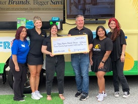  A group of five people in a black shirts and one person in a blue shirt standing in front of a yellow vehicle holding a donation check