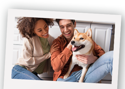 A happy couple, a woman with curly hair and a man with dark hair, sitting on the floor in a cozy kitchen, smiling and hugging a Shiba Inu dog.