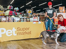 Yellow pet supermarket tablecloth behind a dog available for adoption being pet by a man wearing a red shirt inside of a pet supermarket store